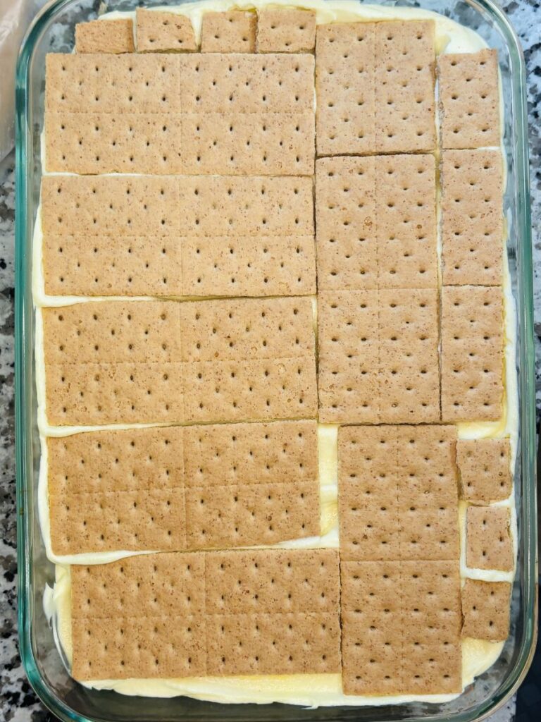 A glass baking dish filled with a dessert topped with a layer of rectangular graham crackers arranged to cover the surface, with creamy filling visible between the cracker layers.