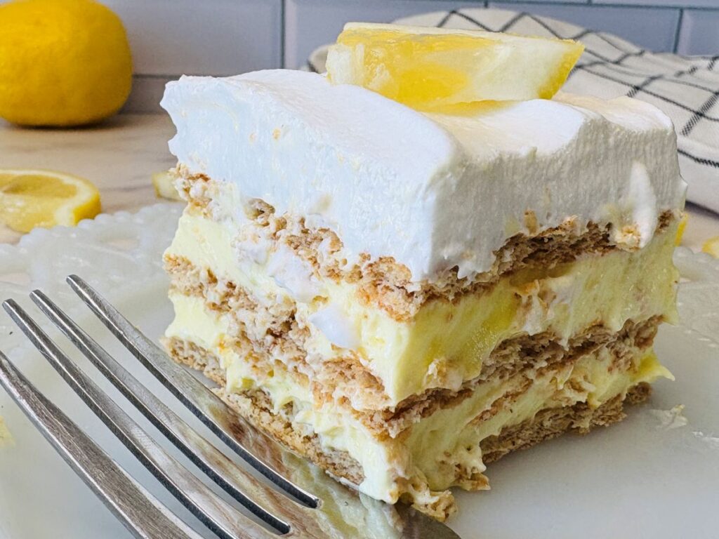 A close-up of a layered lemon dessert with graham crackers, creamy filling, and whipped topping, garnished with a lemon wedge. A fork and a fresh lemon are visible in the background.