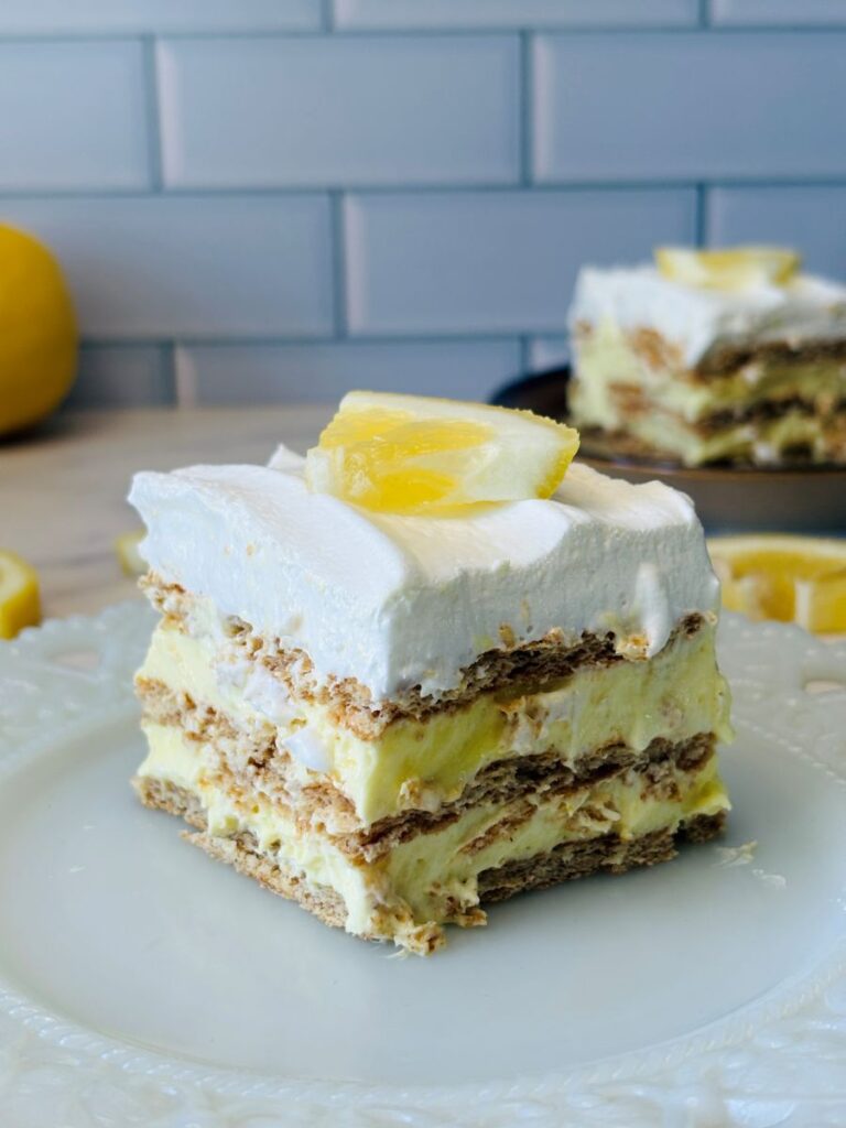 A slice of layered lemon dessert with whipped cream and lemon slices on top, sitting on a white plate; the dessert has visible layers of cream and crackers, with more dessert and a lemon in the background.