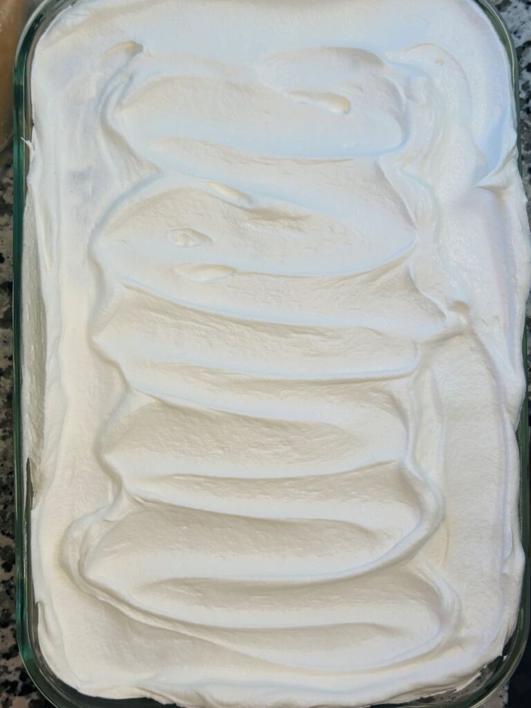 A glass dish filled with a dessert topped with a layer of smooth, swirled whipped cream, sitting on a granite countertop.