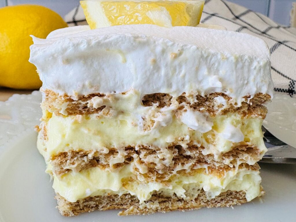 A close-up of a layered lemon dessert with creamy yellow filling, topped with fluffy whipped cream and a slice of lemon, set on a white plate with a lemon and checkered cloth in the background.