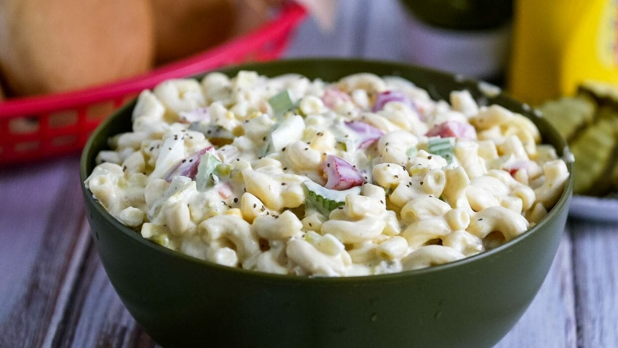 A green bowl filled with creamy macaroni salad, garnished with chopped celery and red onions, sits on a wooden table with a basket of buns and pickles blurred in the background.