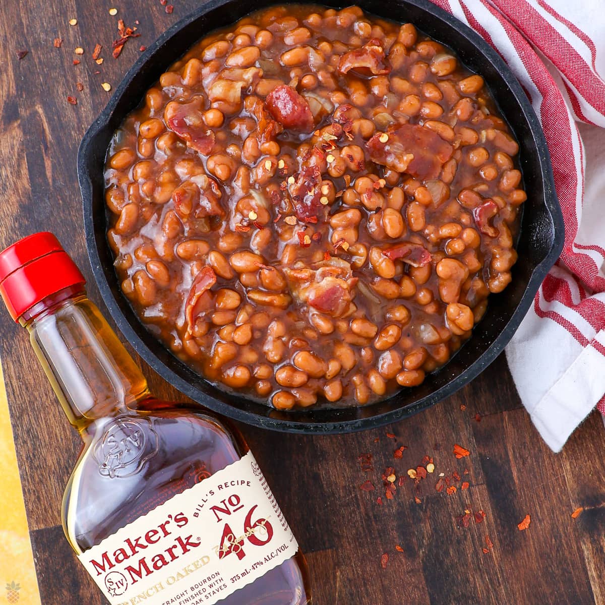 A skillet of baked beans with bacon sits beside a bottle of Maker’s Mark whisky and a red-striped kitchen towel on a wooden table.