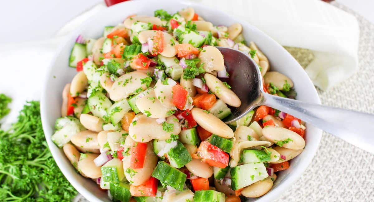 A bowl of colorful bean salad with chopped cucumbers, red bell peppers, onions, and parsley, with a serving spoon resting inside. The salad appears fresh and vibrant, served on a light surface.