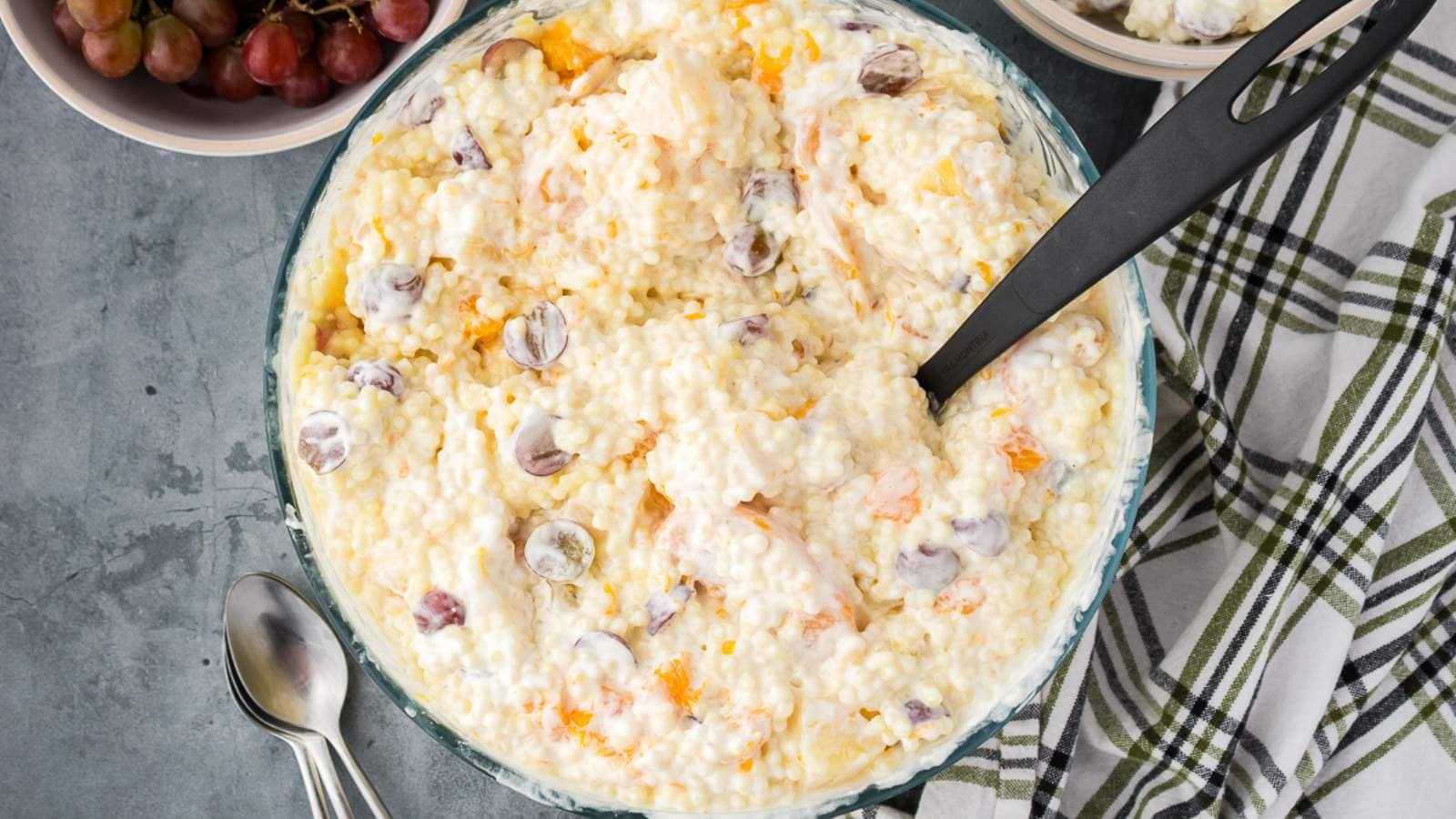 A bowl of creamy fruit salad with grapes and orange pieces, mixed with a whipped topping, sits on a gray surface next to two spoons, a bowl of grapes, and a plaid napkin.