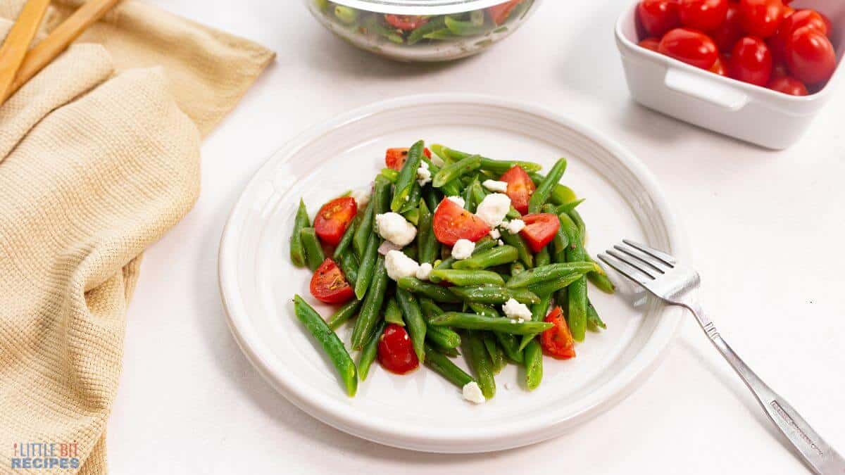 A white plate with green bean salad topped with cherry tomato halves and crumbled feta cheese, placed beside a fork. Nearby are a bowl of salad, a container of cherry tomatoes, and a beige napkin.
