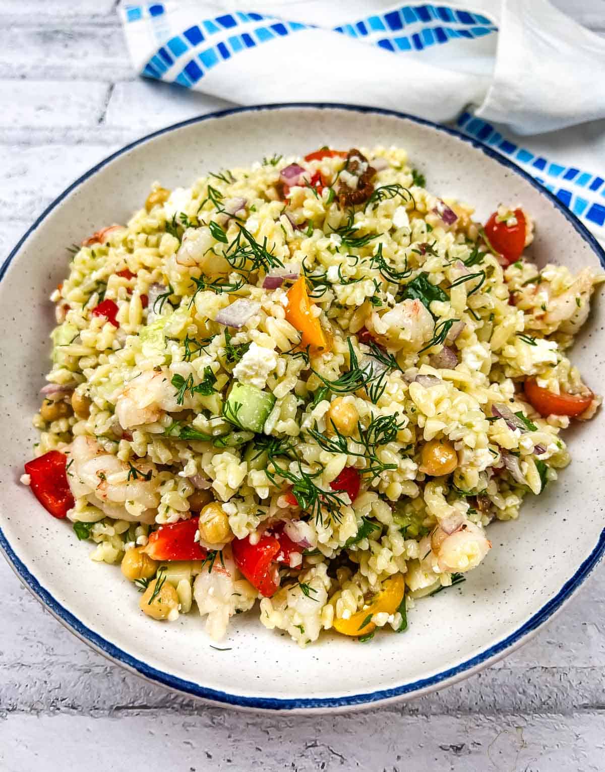 A bowl of bulgur salad mixed with shrimp, cherry tomatoes, chickpeas, red onions, cucumbers, and fresh dill, served on a white plate with a blue rim, placed on a light surface with a blue and white cloth in the background.