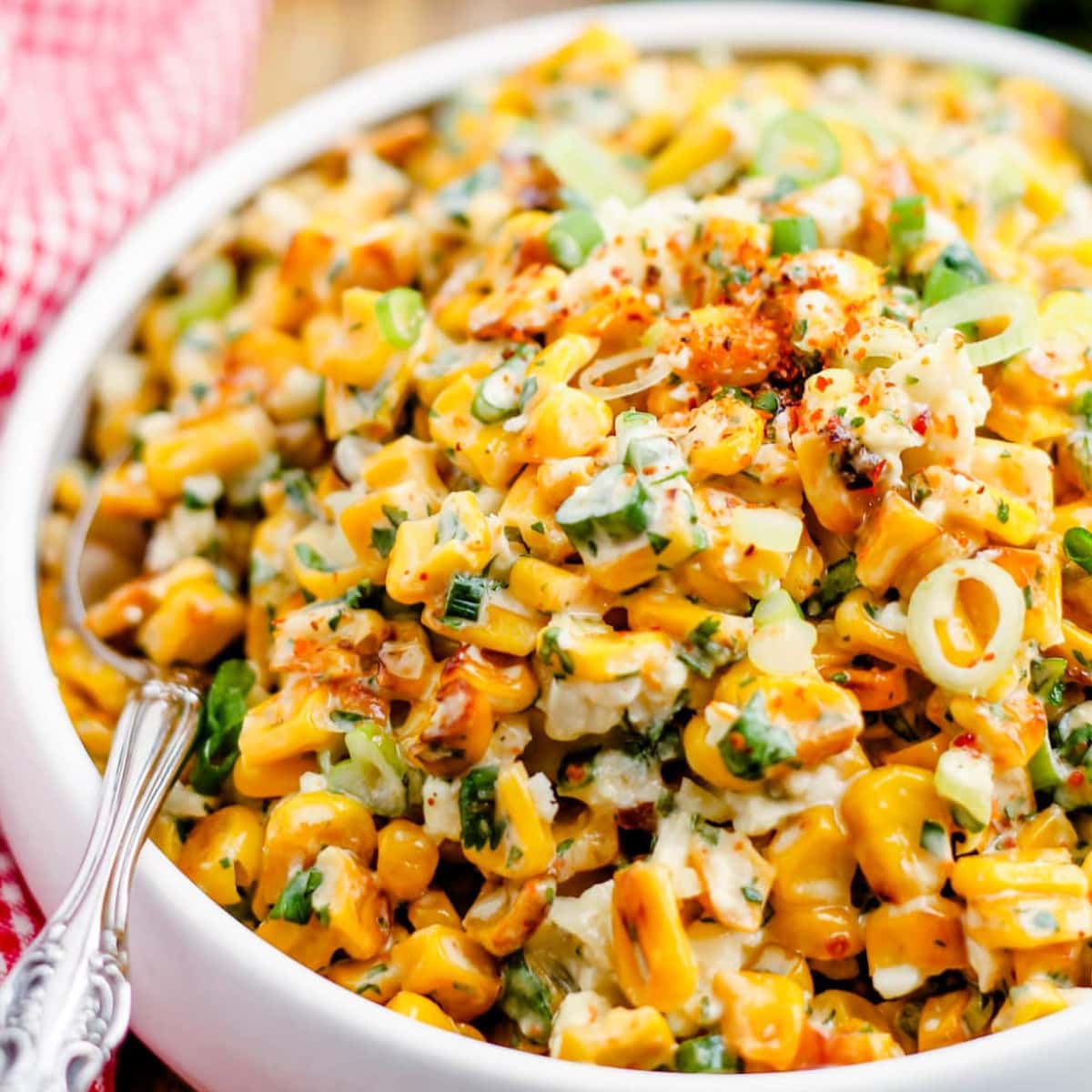 A close-up of a bowl filled with creamy Mexican street corn salad, featuring charred corn, chopped green onions, cilantro, and red chili flakes, mixed with a creamy sauce. A spoon is placed in the bowl.