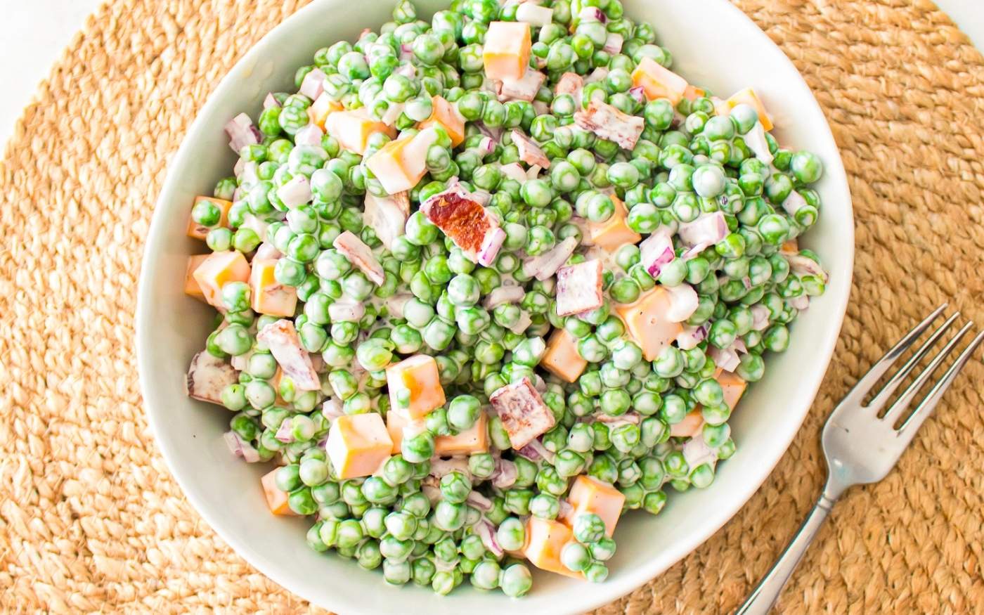 A white bowl filled with creamy pea salad featuring green peas, diced cheese, bacon bits, and red onion sits on a woven placemat next to a fork.