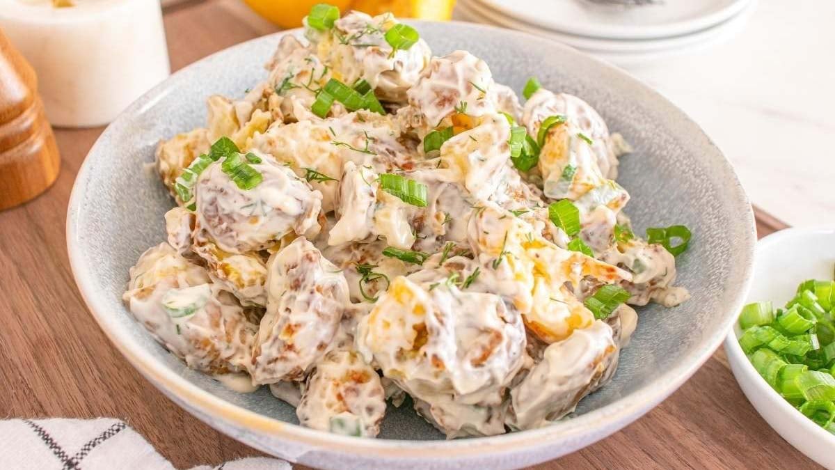 A bowl of creamy potato salad garnished with chopped green onions, served on a wooden surface beside a small dish of extra green onions.