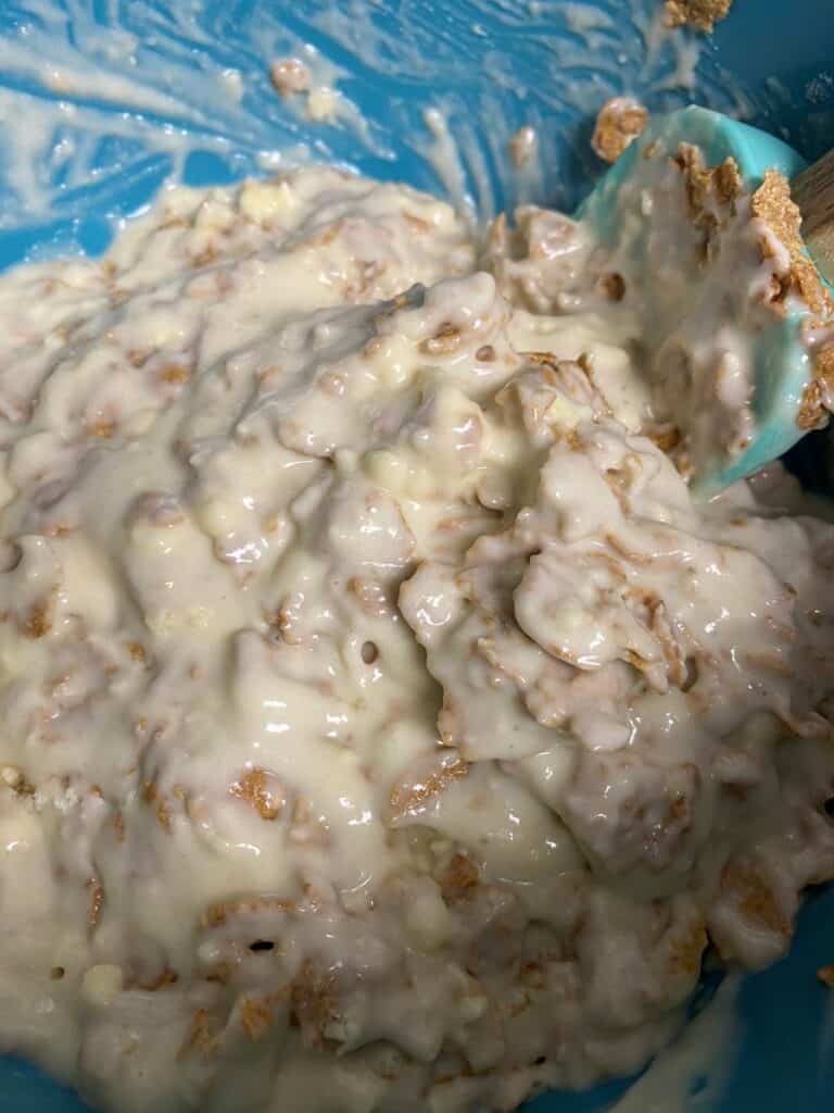 A close-up of a blue mixing bowl filled with a thick, creamy mixture containing bran and batter, being stirred with a blue spatula.