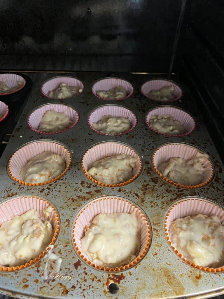 A muffin tin filled with pink paper liners, each holding unbaked, lumpy muffin batter, sits inside an oven. The metal tin is stained and shows signs of frequent use.