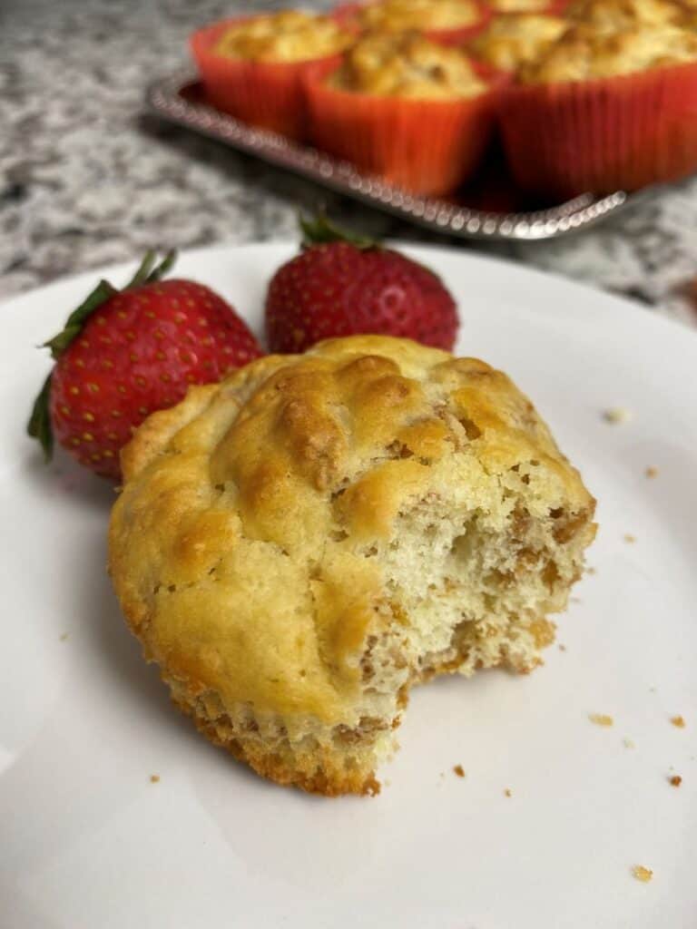 A partially eaten muffin sits on a white plate next to two fresh strawberries, with more muffins in red paper liners on a tray in the background.