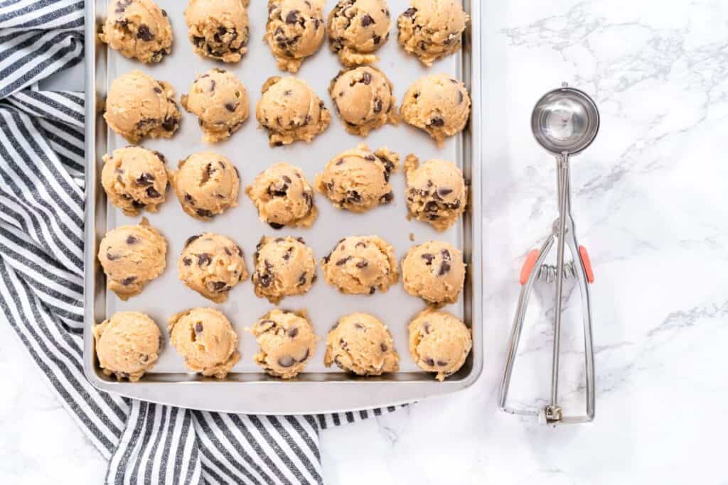 A baking sheet with rows of chocolate chip cookie dough balls on parchment paper, next to a metal cookie scoop and a black-and-white striped kitchen towel on a marble surface.