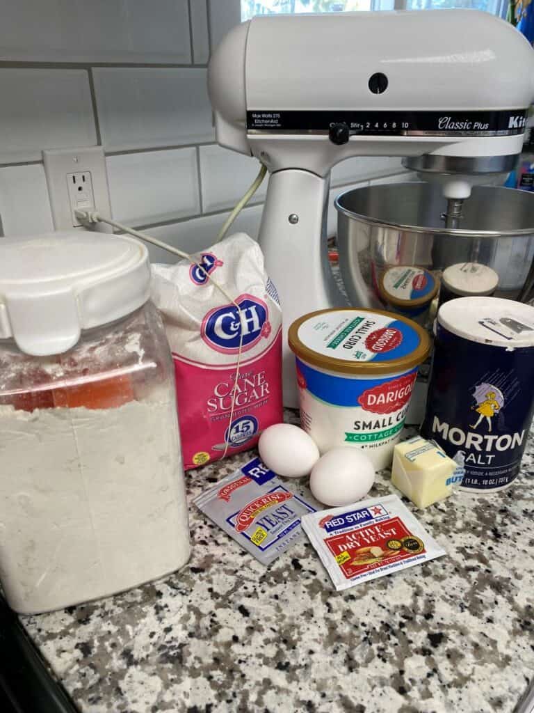 Baking ingredients on a kitchen counter, including flour, sugar, cottage cheese, salt, yeast packets, two eggs, a stick of butter, and a stand mixer in the background.