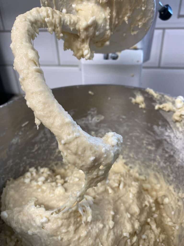 Close-up of a stand mixer with a paddle attachment covered in thick, lumpy batter. The mixing bowl below contains more of the same batter, and white tiled walls are visible in the background.