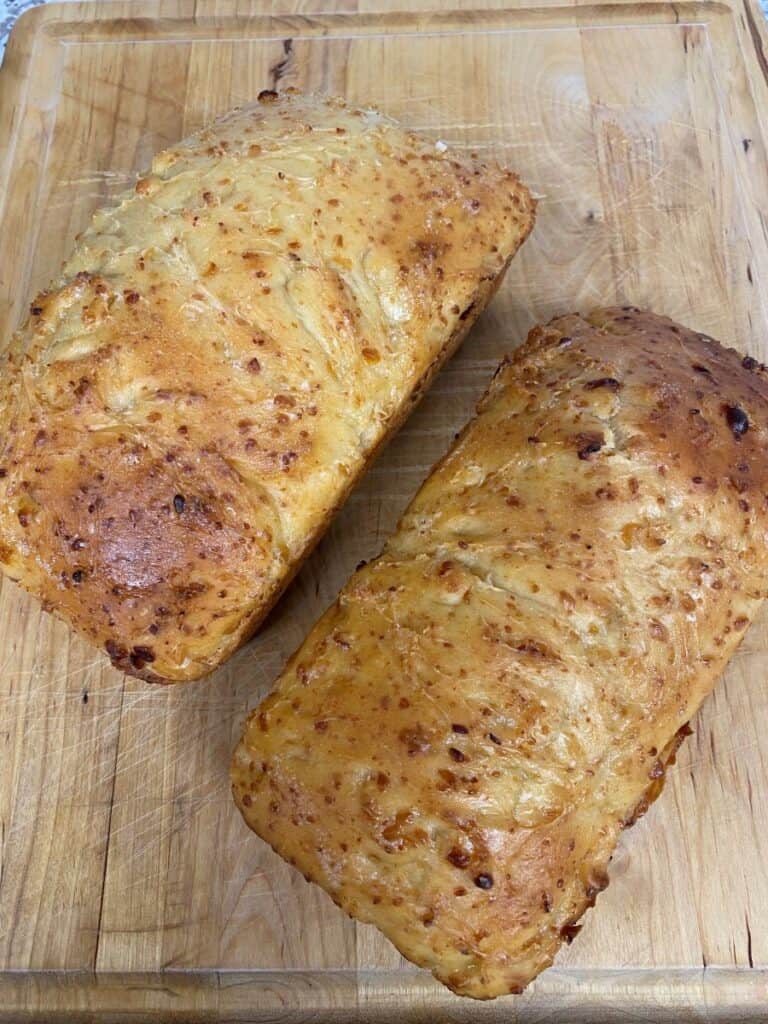Two golden-brown loaves of homemade bread with a slightly glossy crust rest on a wooden cutting board. The loaves have a rustic appearance with visible cheese bits throughout.
