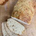 A loaf of homemade bread with a golden crust on a wooden cutting board, partially sliced to reveal a soft, airy interior. Three slices are laid out in front of the loaf.