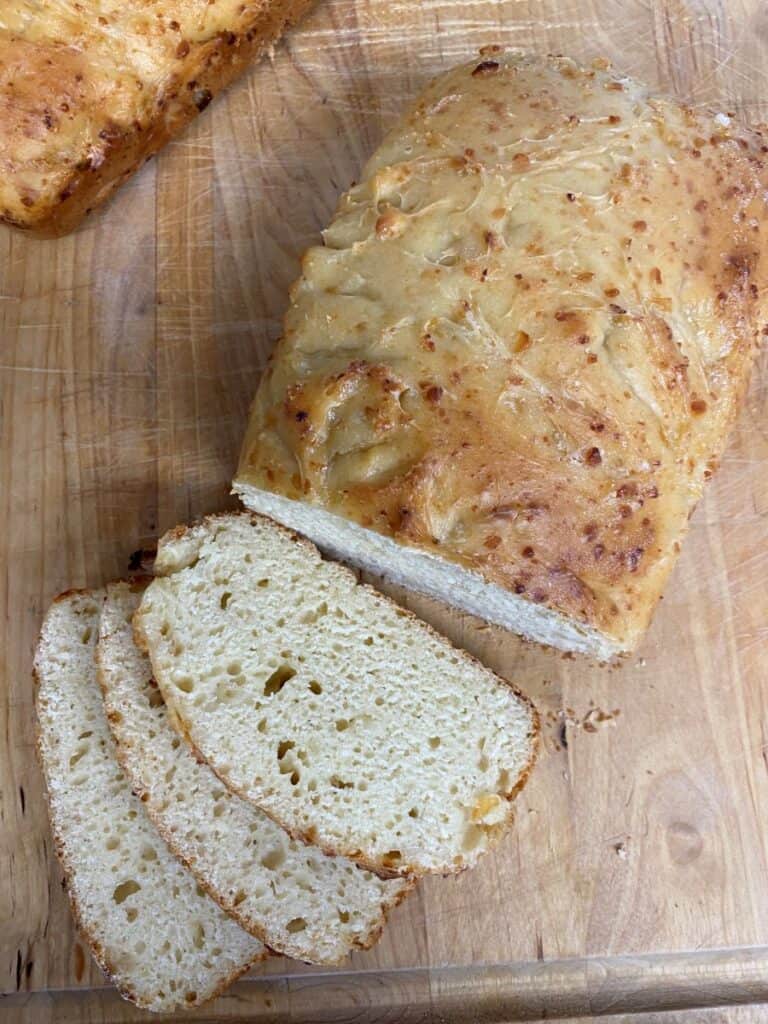A loaf of homemade bread with a golden crust on a wooden cutting board, partially sliced to reveal a soft, airy interior. Three slices are laid out in front of the loaf.