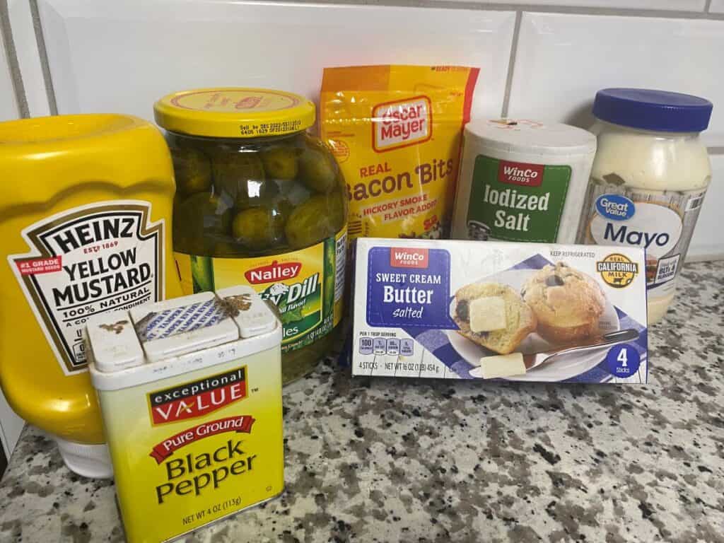 Several condiments and ingredients on a granite countertop, including yellow mustard, dill pickles, bacon bits, salt, mayonnaise, black pepper, and a box of sweet cream butter.
