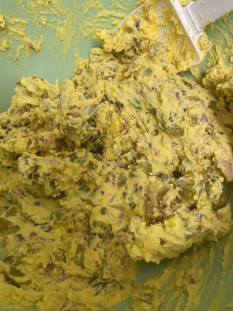 A close-up of a mixture with a creamy yellow base and chunks of green and reddish ingredients, being mixed in a bowl with a white spatula.