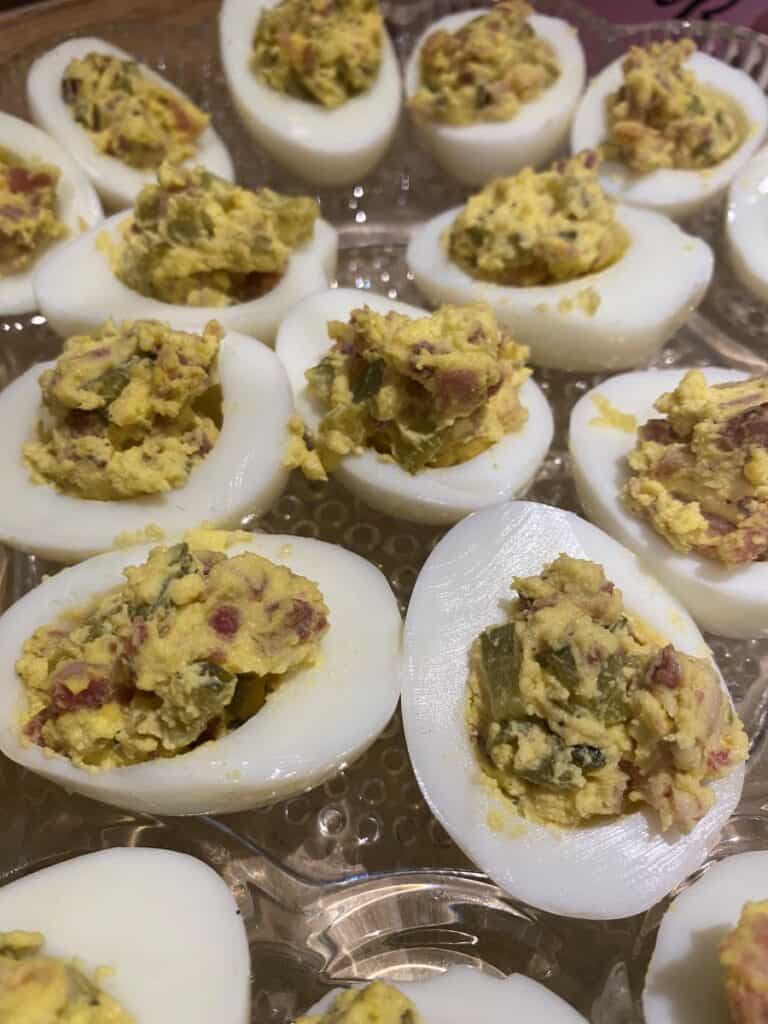 Deviled eggs arranged on a clear platter. The eggs are halved, with a creamy, yellow filling mixed with bits of bacon and herbs placed in the center of each egg white.