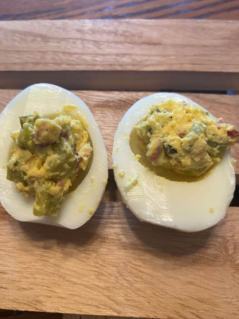 Two deviled egg halves on a wooden surface, each filled with a yellow mixture containing small green and pink pieces, likely chopped vegetables or pickles, with some filling slightly spilling onto the egg whites.