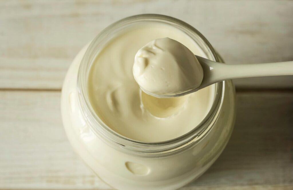 A close-up of a jar filled with creamy white mayonnaise, with a spoon resting above it holding a dollop of the mayonnaise. The background is a light wooden surface.
