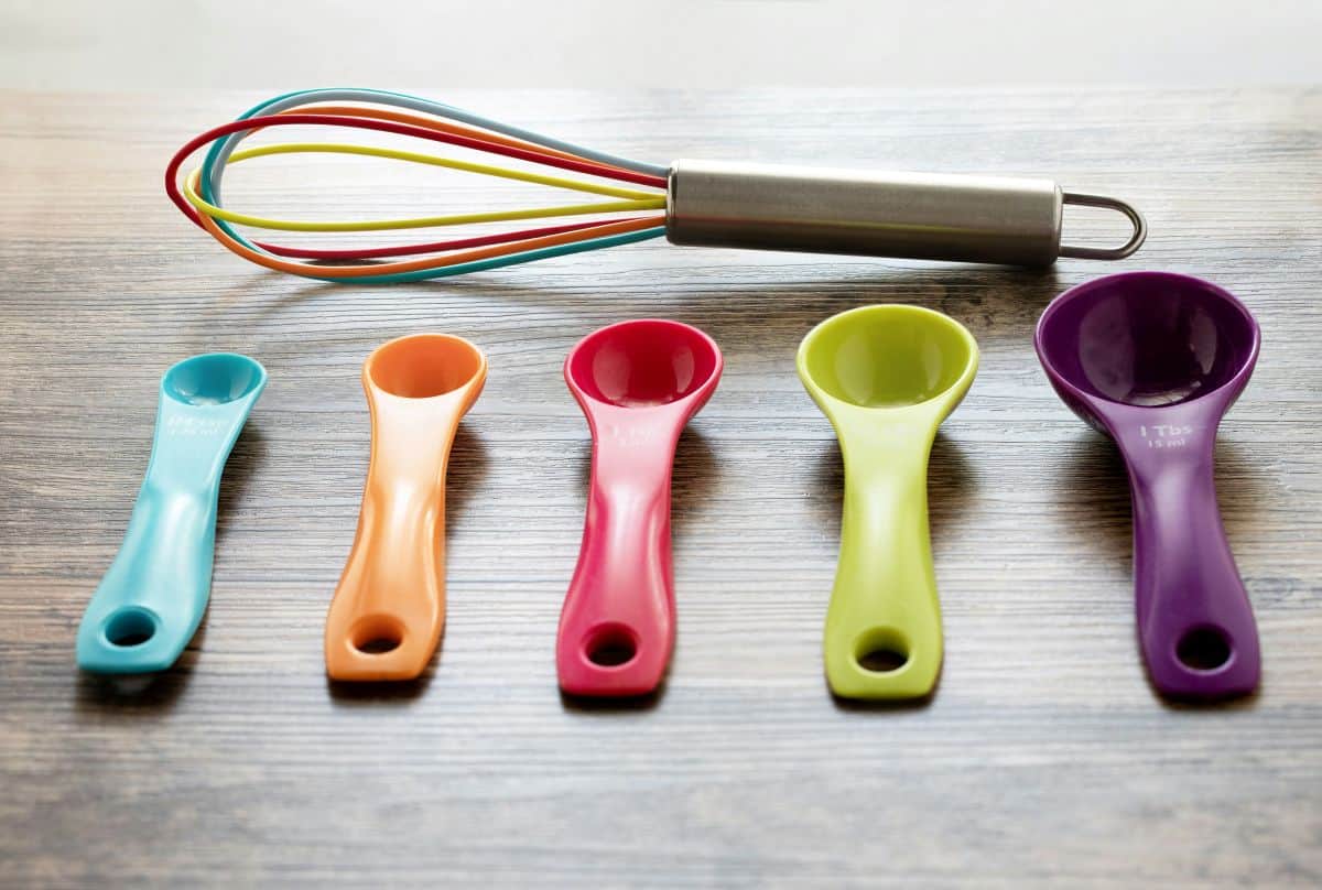A colorful whisk and five measuring spoons in blue, orange, red, green, and purple are arranged on a wooden surface.