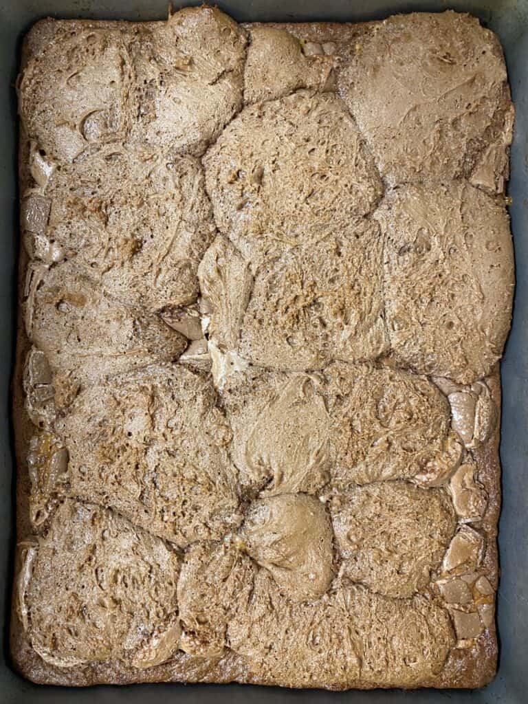 A close-up of a baked, rectangular tray of brown cinnamon rolls or sweet rolls, showing their textured, slightly cracked, and glossy surface, tightly packed together.