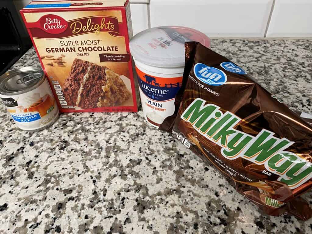 A can of sweetened condensed milk, a box of Betty Crocker German chocolate cake mix, a container of Lucerne plain yogurt, and a bag of Milky Way fun size bars on a granite countertop.