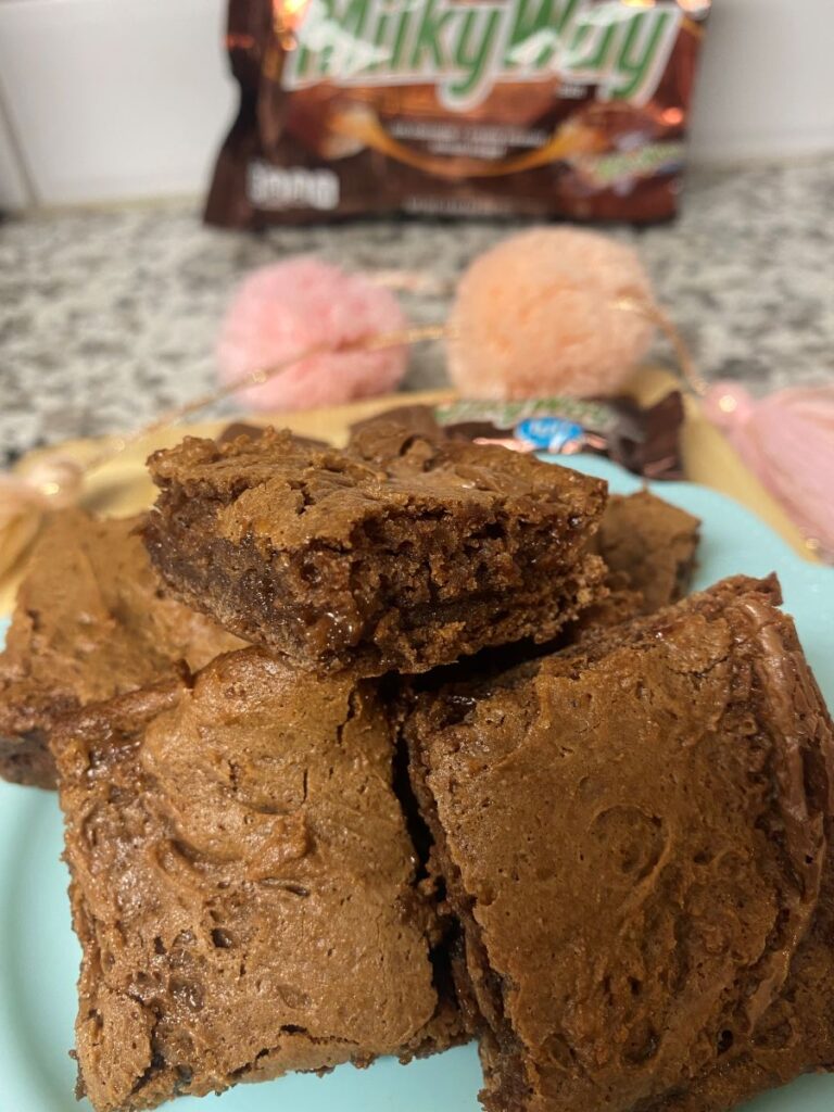 A plate of rich, fudgy chocolate brownies is stacked, with one brownie showing a gooey center. In the background, a Milky Way candy bag is slightly out of focus.
