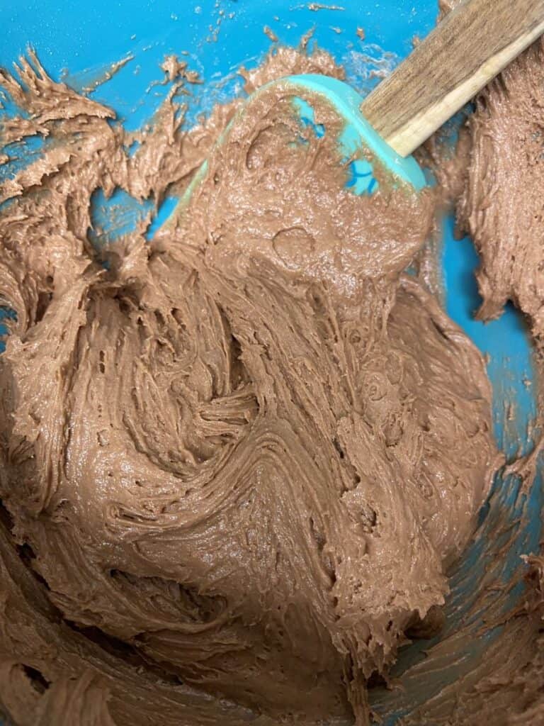 Chocolate cake batter being mixed in a blue bowl with a green spatula. The batter appears thick and creamy, with swirls showing the mixing process.
