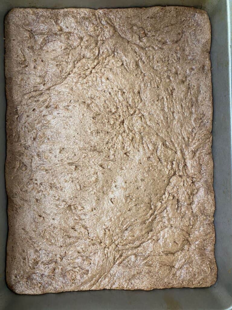 A close-up of a rectangular, golden-brown baked dessert in a metal pan, possibly a tray of brownies or blondies, with a slightly uneven, textured surface.