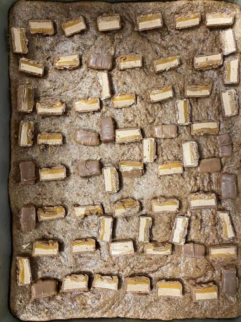 A baking tray filled with unbaked cookie dough topped with evenly spaced pieces of chopped chocolate and caramel candy bars.