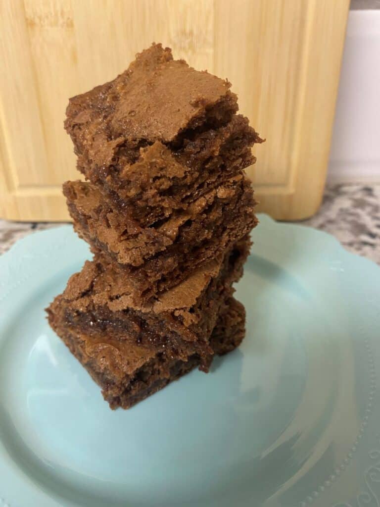 A stack of four fudgy chocolate brownies sits on a light blue plate, with a wooden cutting board in the background. The brownies have a cracked, chewy top and a rich, dense interior.