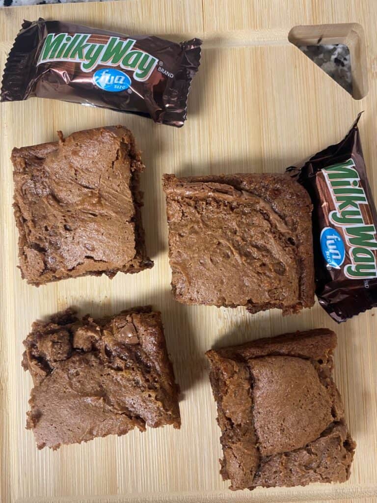 Four chocolate brownies are placed on a wooden surface, with two unwrapped Milky Way Fun Size chocolate bars nearby. The brownies appear thick and fudgy.