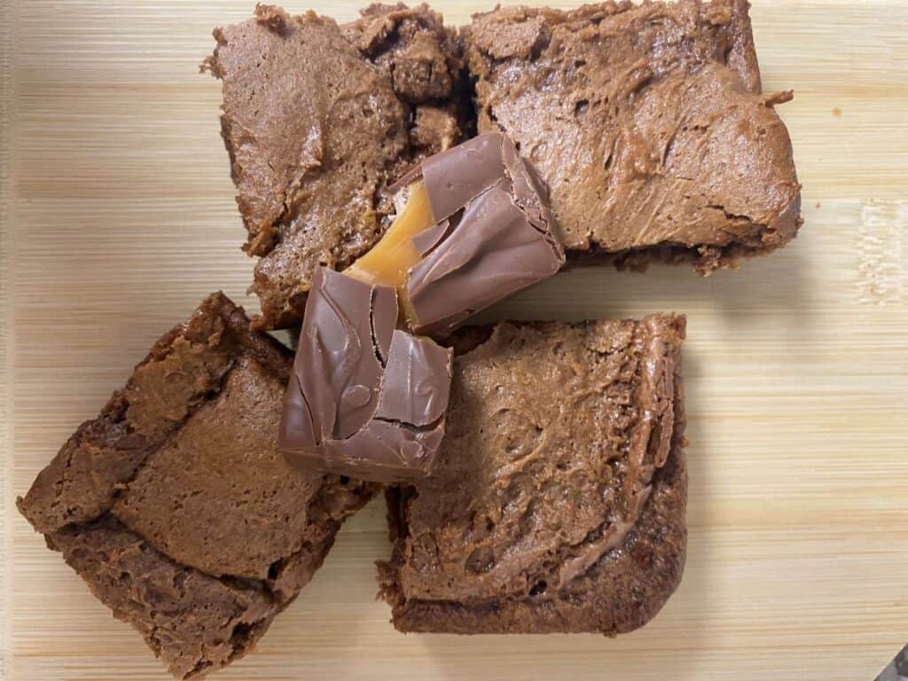 Three chocolate brownies arranged on a light wooden surface with a piece of chocolate-covered caramel candy placed on top of the brownies.