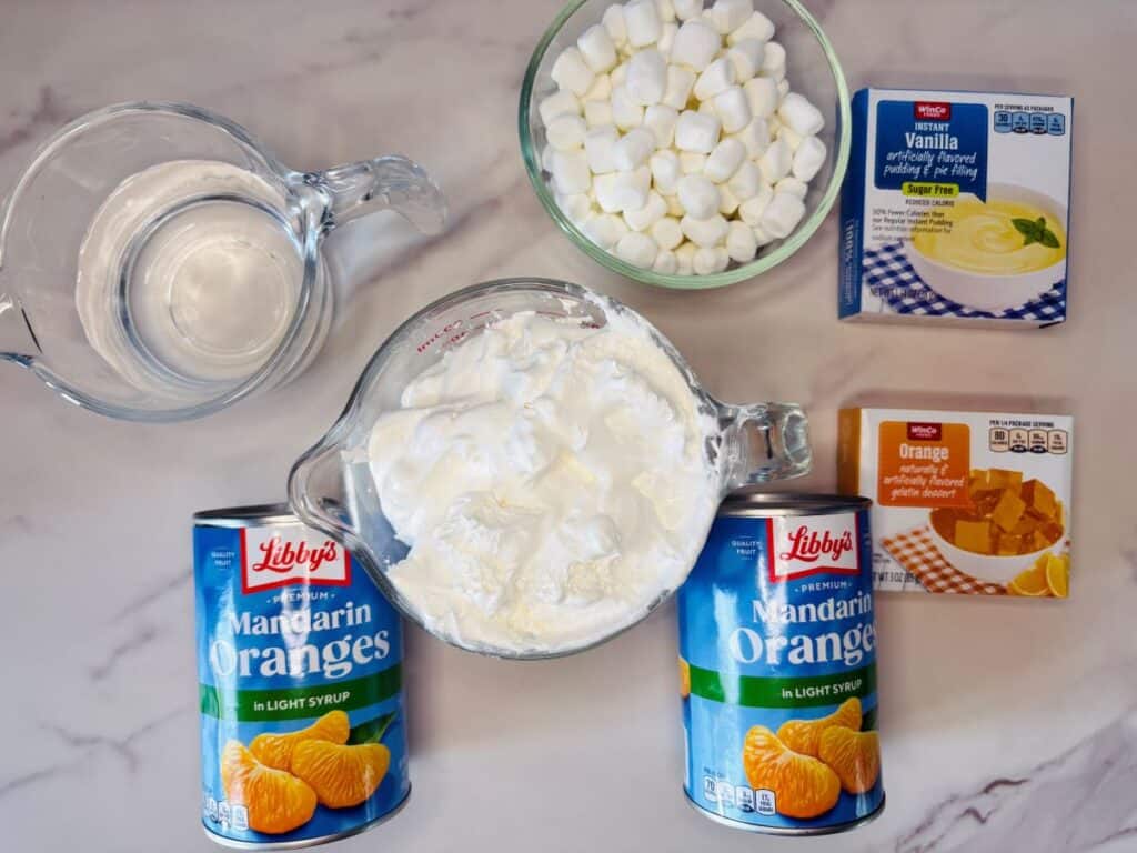 Two cans of mandarin oranges, a measuring cup of whipped topping, a bowl of mini marshmallows, two boxes of gelatin dessert vanilla and orange flavors, and a pitcher of water arranged on a marble surface.