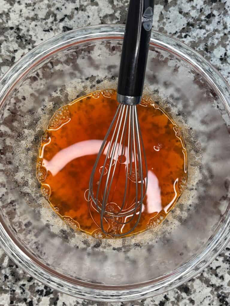 A metal whisk rests in a clear glass bowl filled with a reddish-orange liquid mixture, sitting on a speckled gray countertop.