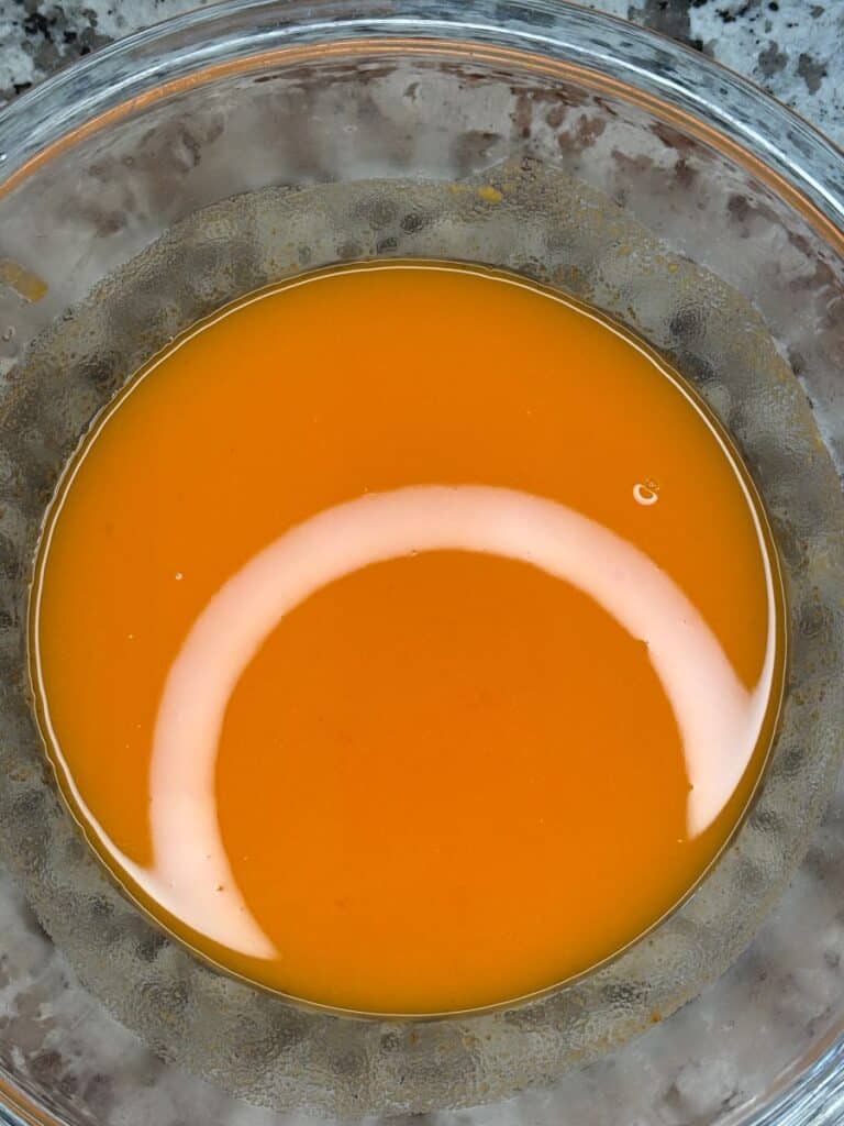 A clear glass bowl filled with bright orange liquid sits on a gray, speckled surface. A reflection of light forms a white arc across the surface of the liquid.