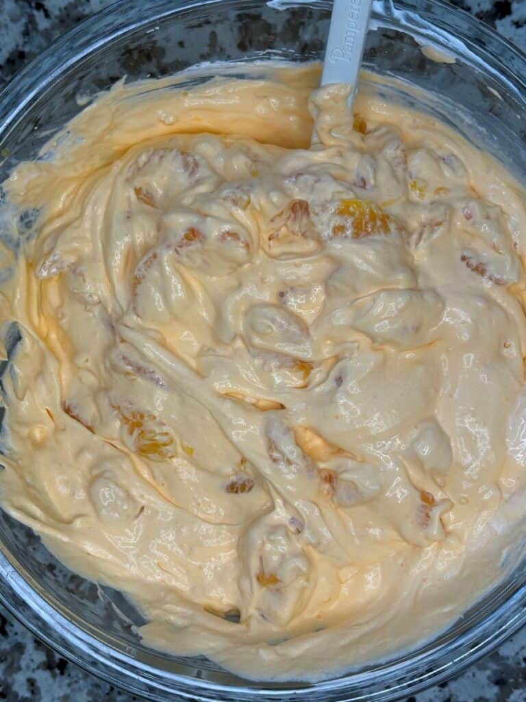 A glass bowl filled with creamy orange fruit salad, mixed with chunks of fruit and a spoon resting inside. The mixture looks thick and fluffy.