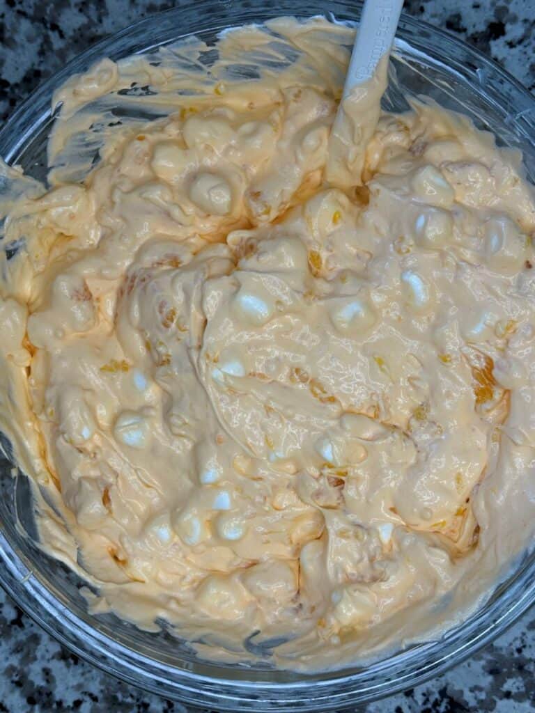 A glass bowl filled with a creamy orange mixture that contains small white marshmallows and bits of fruit, with a spoon partially visible in the mixture. The bowl is on a granite countertop.