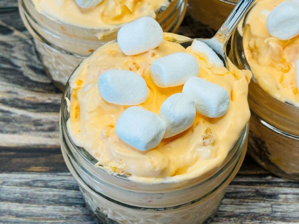 A small glass jar filled with creamy orange fluff dessert, topped with several mini marshmallows, sits on a wooden surface. Other jars of the same dessert are partially visible in the background.