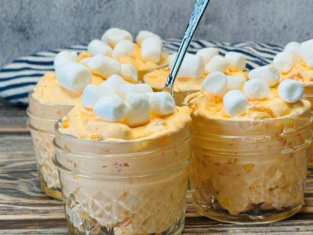 Four small glass jars filled with creamy orange dessert topped with mini marshmallows are arranged on a rustic wooden surface, with a spoon in one jar and a striped cloth in the background.