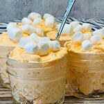 Four small glass jars filled with creamy orange dessert topped with mini marshmallows are arranged on a rustic wooden surface, with a spoon in one jar and a striped cloth in the background.