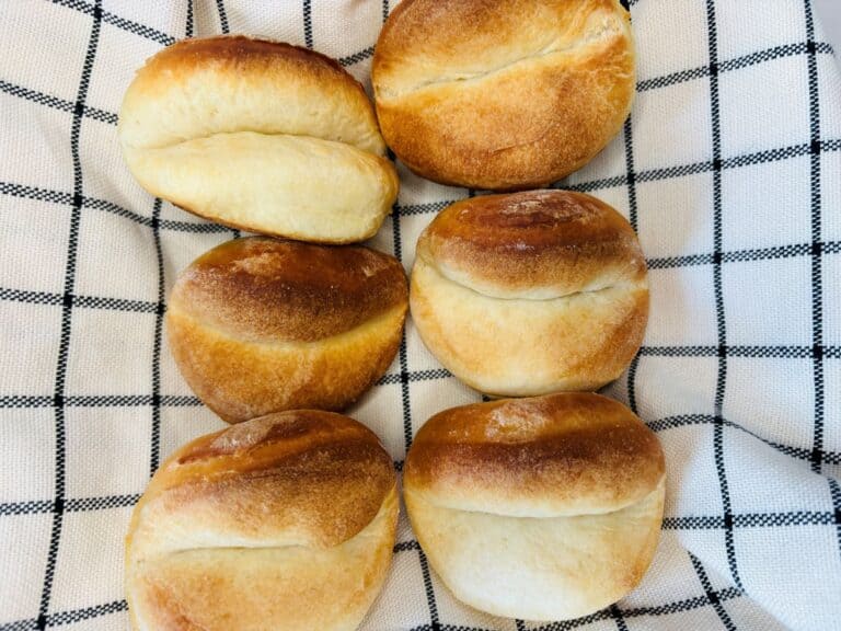 Six round, golden-brown bread rolls are arranged on a white cloth with a black grid pattern. The rolls have a slightly split top and a soft, baked texture.