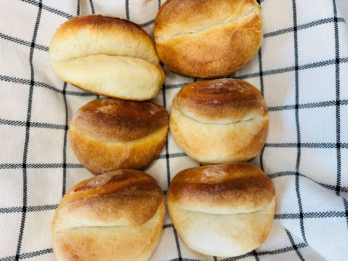 Six round, golden-brown bread rolls are arranged on a white cloth with a black grid pattern. The rolls have a slightly split top and a soft, baked texture.