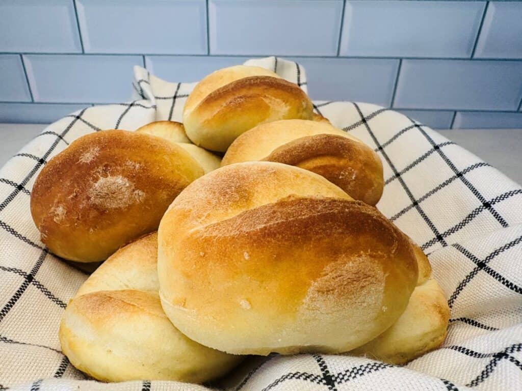 A basket lined with a white and black checkered cloth holds several golden-brown, freshly baked bread rolls in front of a white tiled wall.