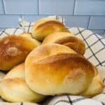 A basket lined with a white and black checkered cloth holds several golden-brown, freshly baked bread rolls in front of a white tiled wall.